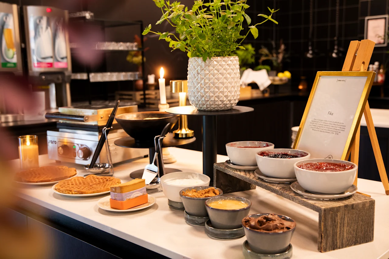 Et koselig fika-oppsett med ferske vafler og diverse deilige pålegg, inkludert syltetøy og sjokoladepålegg, med et Fika-skilt.