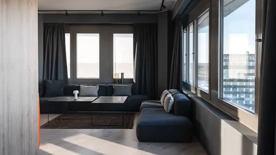 A modern living room features a black sofa with cushions facing large windows. A low coffee table holds a small lamp.