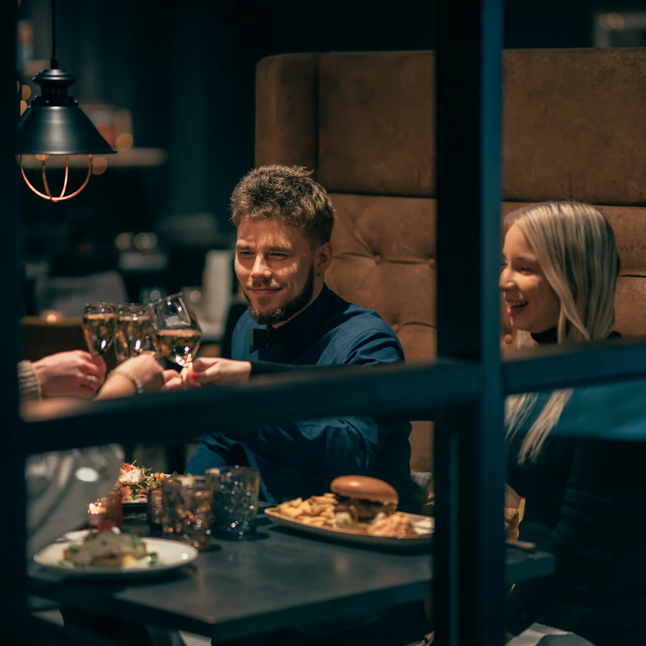 A group of people raise glasses, smiling, seated in a cozy restaurant booth with plates of food like burgers, fries, and drinks on the table.