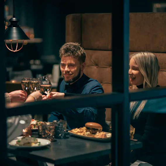 Ryhmä ihmisiä nostaa laseja, hymyillen, istuen kodikkaassa ravintolakojussa lautasilla ruokaa, kuten hampurilaisia, ranskalaisia ja juomia pöydällä.