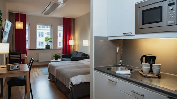 A small studio apartment includes a kitchen with a microwave, sink, and kettle. Adjacent is a living area with a bed, table, chairs, sofa, and lamp, near large windows.