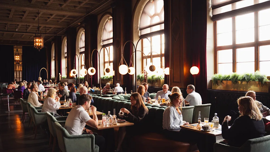 Personer äter och samtalar i en rymlig, elegant designad restaurang med höga fönster, moderna cirkulära lampor och frodig grönska på fönsterbrädorna. Atmosfären är livlig och välkomnande.