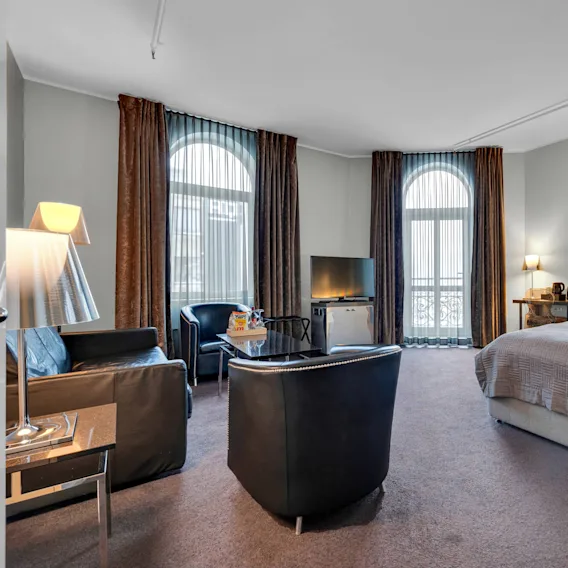 Et hotellrom med seng, lenestoler og TV. Store vinduer med gardiner slipper inn naturlig lys. Et lite bord har lampe og kaffemaskin.