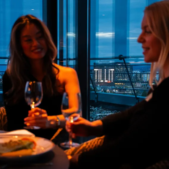 To personer skåler med vinglass ved et restaurantbord foran store vinduer om kvelden med bylys i bakgrunnen.