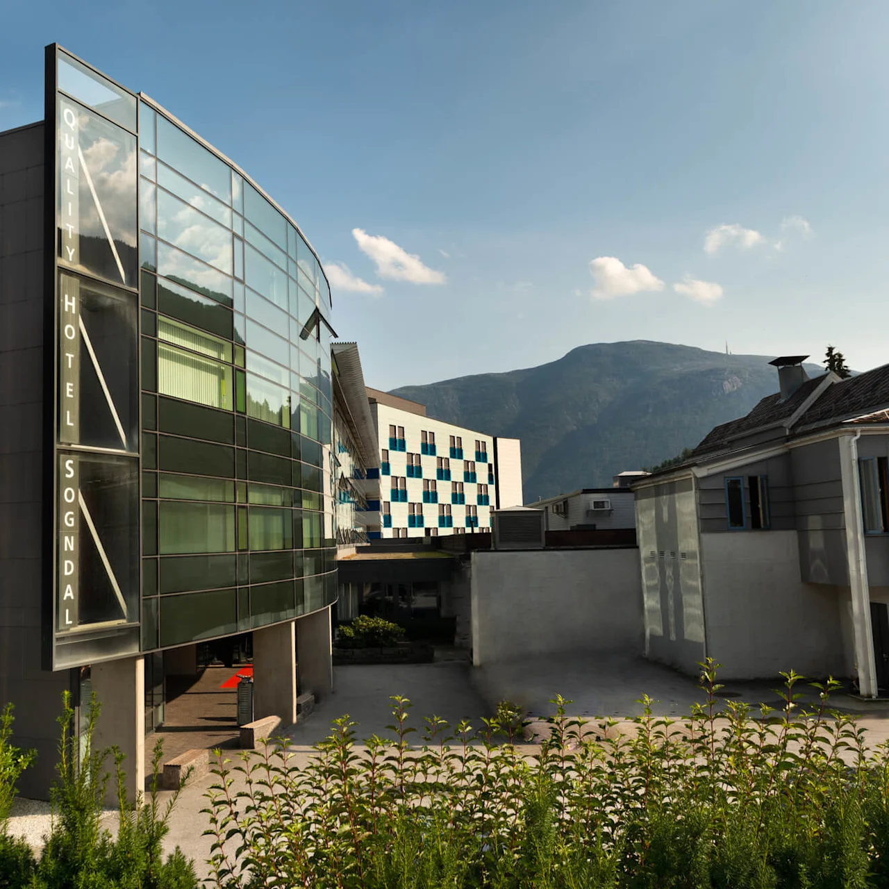 En modern glasbyggnad märkt "QUALITY HOTEL SOGNDAL" står bredvid traditionella byggnader, mot en bergig bakgrund under en klarblå himmel.