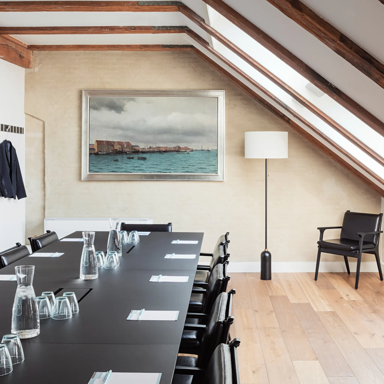 A long black conference table is set with glasses, carafes, and notepads, positioned under a sloped ceiling with exposed beams. A framed seascape hangs on the wall.