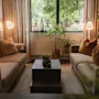 A cozy living room with two sofas, a coffee table, and plants, bathed in natural light from a large window overlooking greenery.
