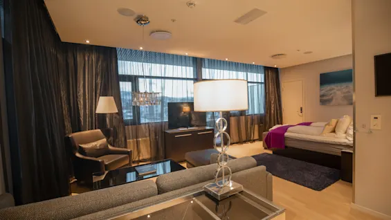 Et moderne hotellrom har en lampe på et bord, lenestol, TV, seng med lilla teppe og dekorativ veggkunst, omgitt av gardiner fra gulv til tak og myk belysning.