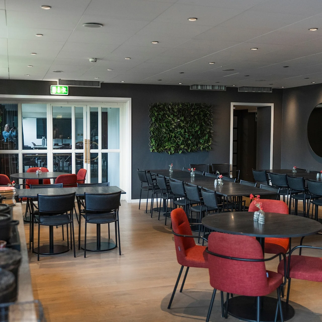 Bord og stolar i rødt og svart är placerade i en modern restaurangmiljö. En hängande växt och en väggmonterad växtlighet gör dekorationer. Text på spegel: "Hello there good looking!"