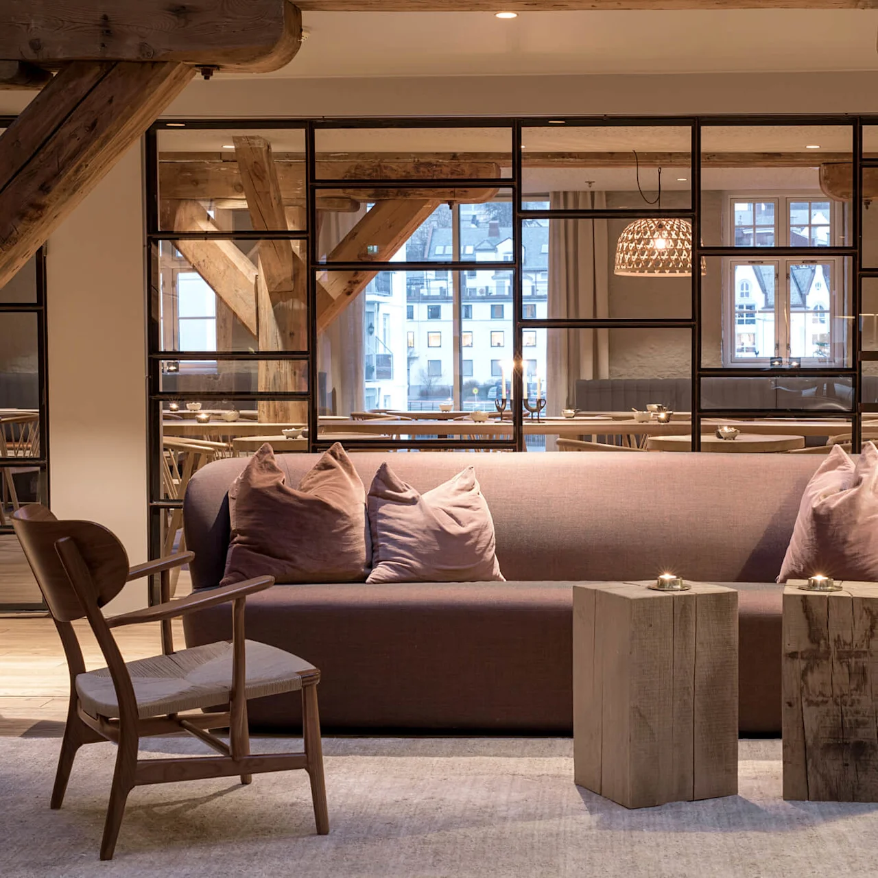 Sofa with cushions sits on a rug, flanked by wooden tables and a lamp. Background features large windows and wooden beams, with visible dining area.