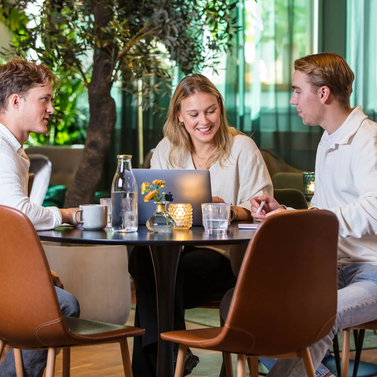 Three people are engaged in a conversation at a table with a laptop, in a cozy indoor setting with greenery.