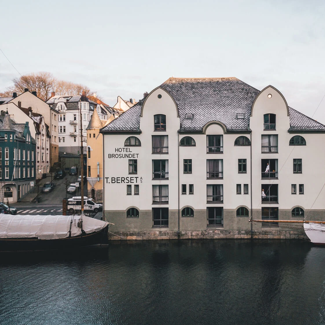 A white building labeled "Hotel Brosundet T. Berset AS" stands by a calm waterfront, surrounded by colorful buildings and moored boats under a clear sky.