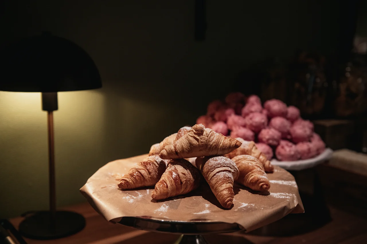 Lähikuva kasasta kullanruskeita croissanteja tomusokerilla, telineellä lampun ja vaaleanpunaisten leivonnaisten kanssa taustalla.