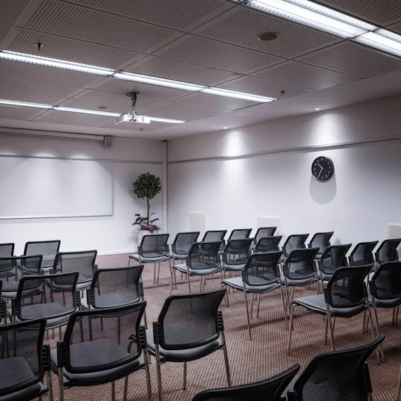 Rows of empty black chairs face a whiteboard in a conference room. A clock on the wall reads four o'clock, and a plant decorates the corner.