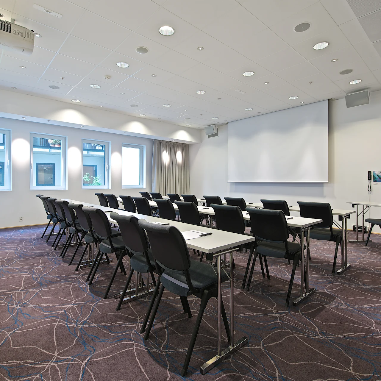 Rader av svarta stolar vända mot bord med vita ytor, belägna i ett väl belyst konferensrum. En projektor och en stor skärm är installerade framtill.