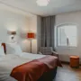 Viihtyisä hotellihuone, jossa on mukava sänky, ikkuna, raidallinen nojatuoli ja pieni työpöytä tuolineen.