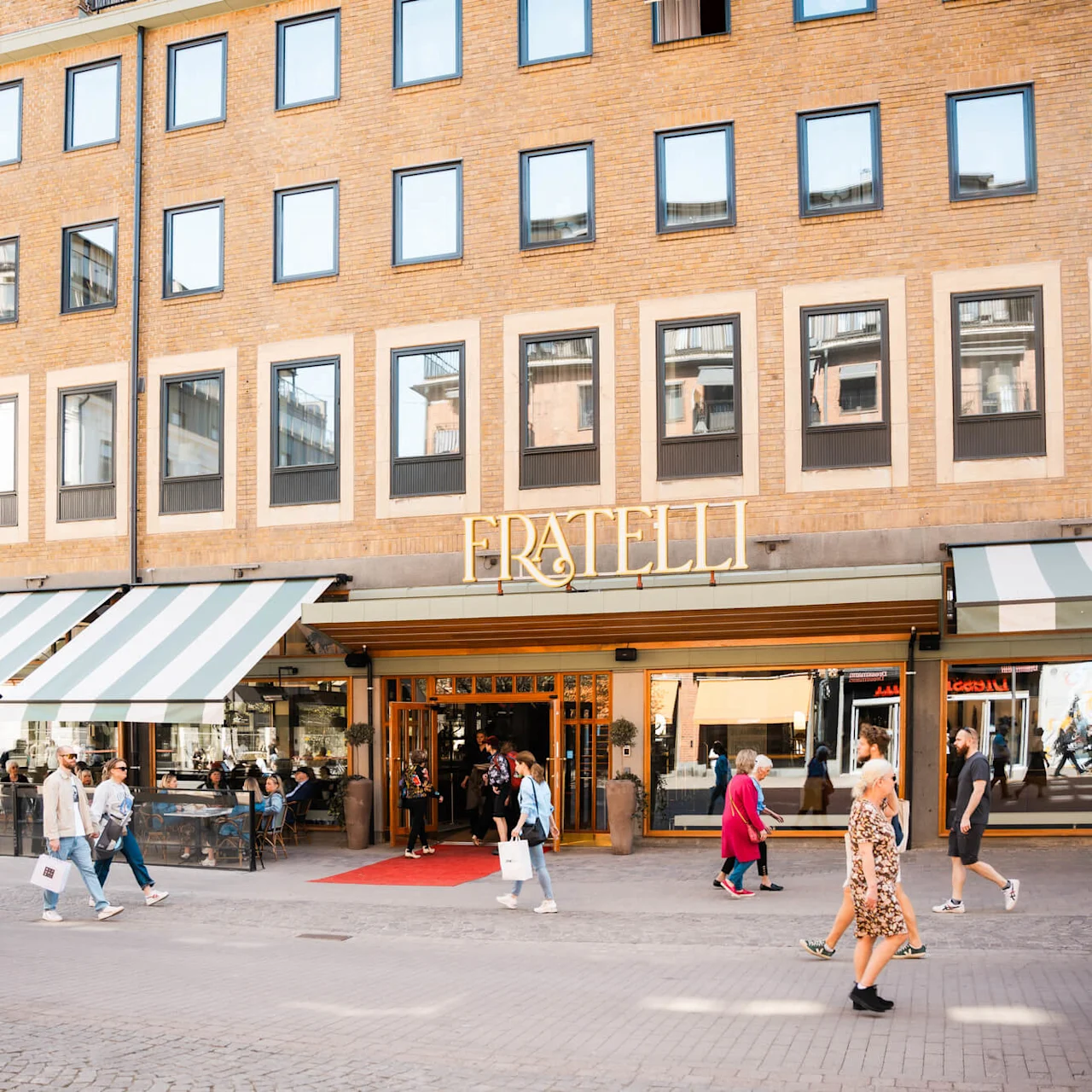 A brick building with windows, where a restaurant named "FRATELLI" has a striped awning. People walk by on a sunny day, with some sitting outside at tables.
