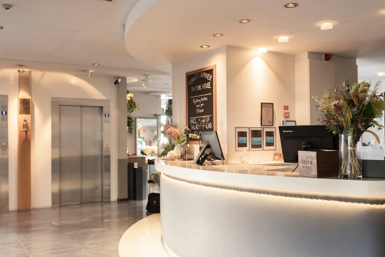 En hotellreception med datorer, blommor och skyltar som säger TONIGHTS DINNER ON THE HOUSE och GREEN MARKET. Hissar syns.