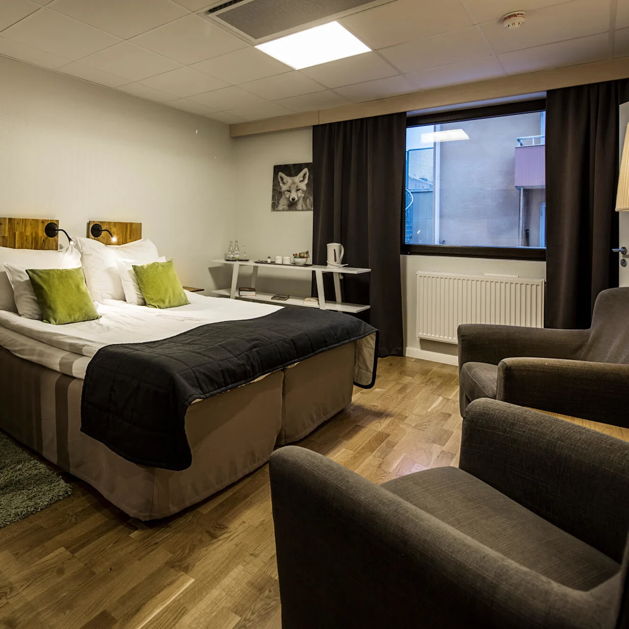 A neatly made bed with green accents in a modern bedroom, two armchairs in the foreground.