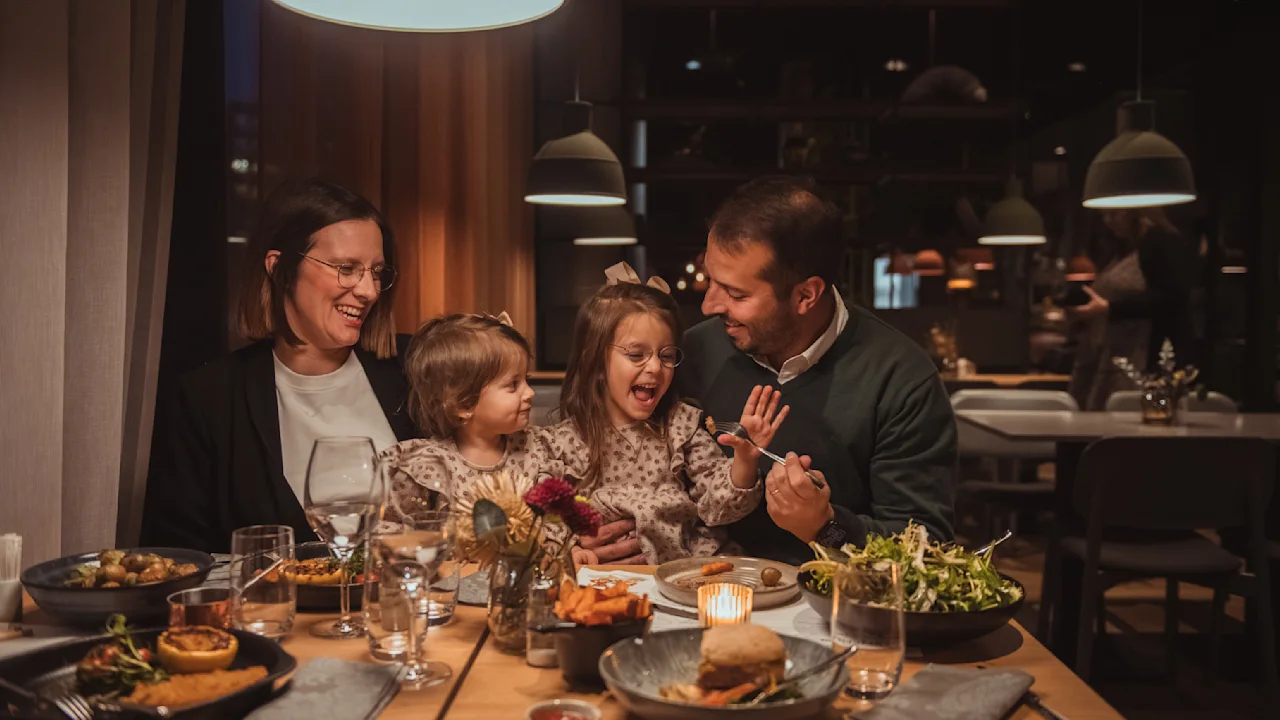 En familie nyder en hyggelig middag på en varmt oplyst restaurant, deler smil og latter. Tallerkener med forskellige retter og nogle glas står på bordet, skaber en glædelig middag.