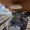 Utomhusterrass med moderna möbler och ljuskronor; har utsikt över järnvägsspår och en flod. Omgiven av stadssilhuett under en delvis molnig himmel.