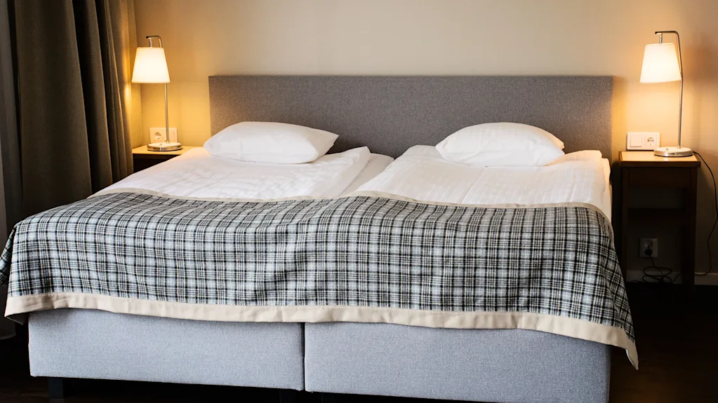 Viihtyisä hotellihuone, jossa kaksi sänkyä, ruudullinen peitto ja yöpöytälamput, valmiina mukavaan oleskeluun.