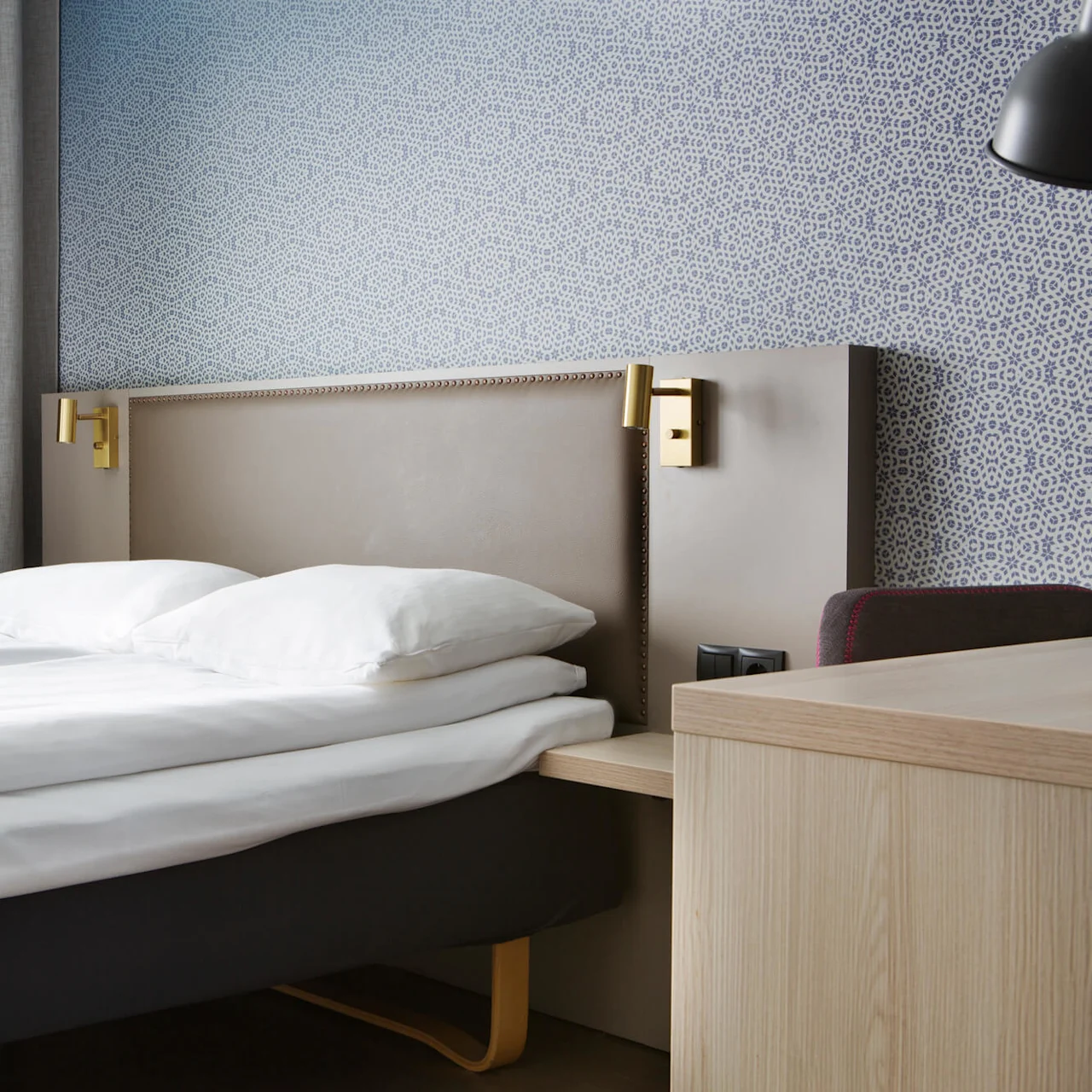 A neatly made bed with white bedding in a room with patterned wallpaper and a window with grey curtains.