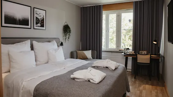 En pent dobbeltseng med to foldede badekåper står i et hotellrom. Et skrivebord med lampe er ved det gardinbelagte vinduet, og innrammet kunst dekorerer veggen.
