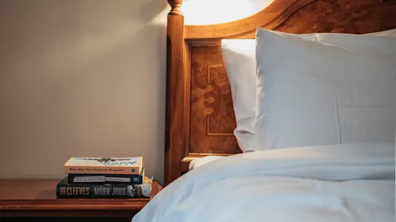 En mysig sängscen med böcker på ett nattduksbord bredvid en säng med vita lakan. Synliga boktitlar: Mork Jord, Jonas Mostrom, Ann Cleeves.