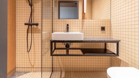Et moderne badeværelse med en rektangulær hvid håndvask på en sort bordplade. Omgivende beige fliser, en glasskærmvæg og et vægspejl fuldfører det minimalistiske design.