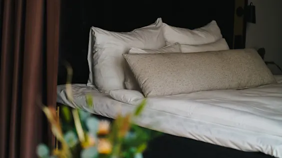 En pænt redt seng med hvide lagner og puder står bag brune gardiner. En sløret buket blomster i forgrunden tilføjer farve til rummet.