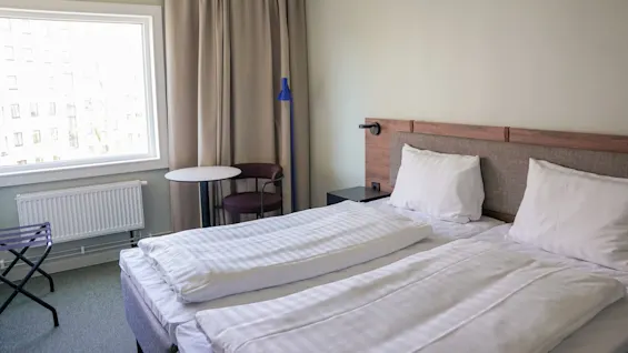 En dobbeltseng med hvitt sengetøy står plassert i et hotellrom. Et vindu med åpne gardiner viser en tilstøtende bygning. Et lite bord med en stol står i nærheten.