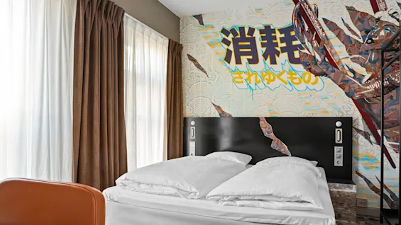 Bed with white linens against a wall featuring colorful Japanese artwork and text. Brown and white curtains filter soft light from large windows.