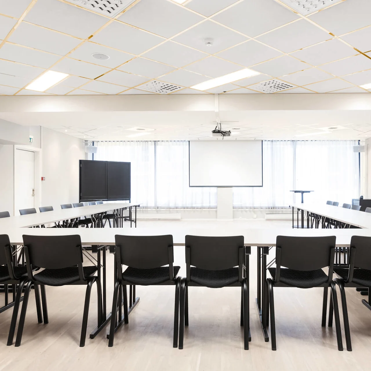 Et konferencelokale har tomme borde og stole arrangeret i en U-form, vendt mod et tomt lærred. Lyse loftlys og store vinduer oplyser rummet.