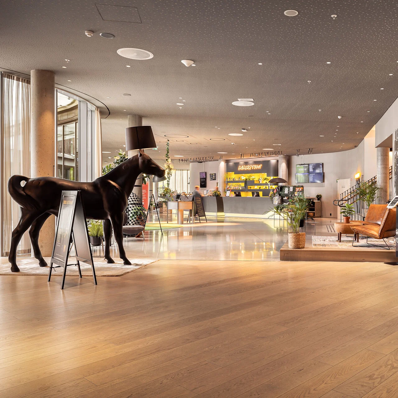 A horse statue with a lampshade head stands near a reception desk in an upscale lobby with modern seating and wooden decor. Sign reads "RECEPTION."