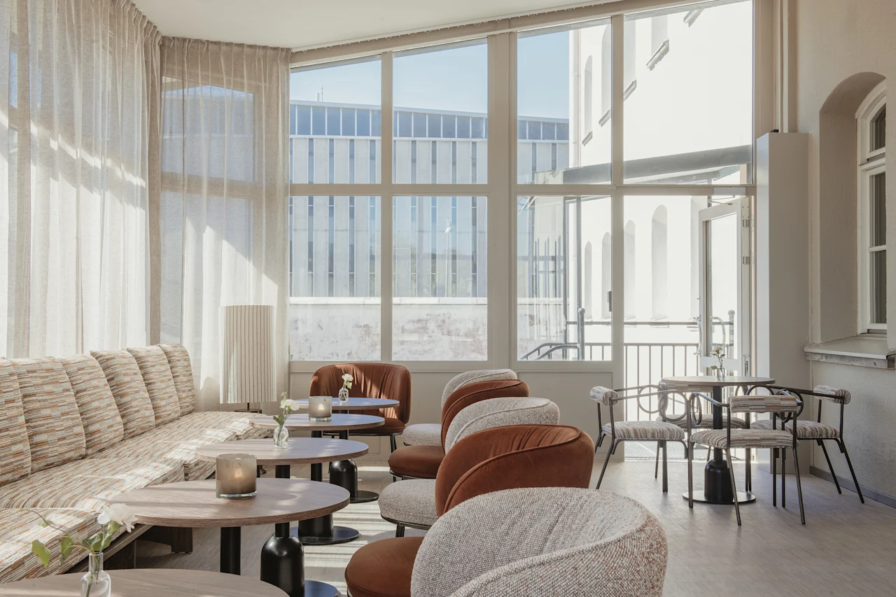 A bright and inviting lounge area with comfortable seating, small tables, and large windows looking out onto an urban scene.