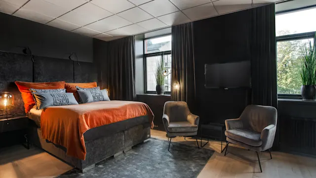 En stor seng med oransje sengetøy står i et moderne, dempet belyst soverom. To grå stoler vender mot en veggmontert TV. Store vinduer med mørke gardiner viser grønt utenfor.