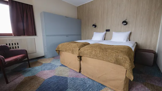 Two neatly made single beds with brown blankets stand in a hotel room. A patterned, colorful carpet covers the floor, while a window with closed curtains and a cushioned chair complete the setting.