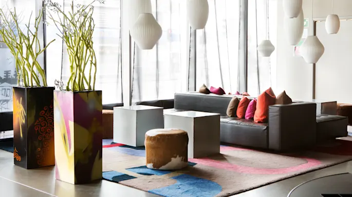 Sofa with colorful pillows, standing still, in a modern room with large windows, vibrant plants, geometric tables, and hanging white lamps.