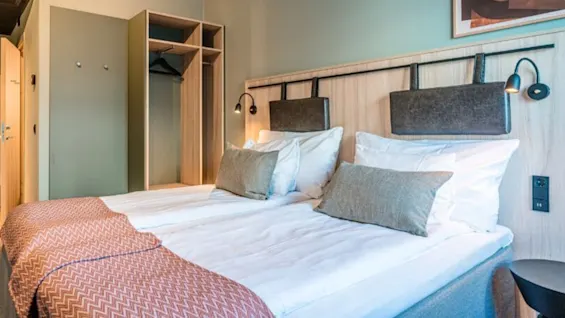 A neatly made double bed with brown cushions and patterned throws stands in a hotel room, accompanied by a wardrobe and nightstand under a well-lit, framed picture.