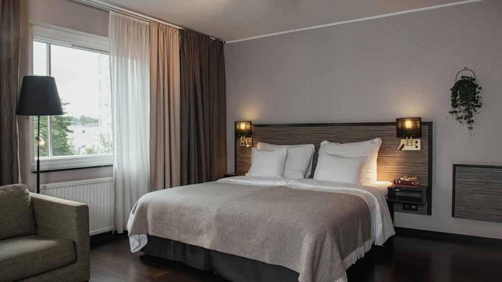 A cozy hotel room with a large bed, a comfortable armchair by the window, and warm lighting, inviting relaxation.