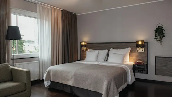 Viihtyisä hotellihuone, jossa on suuri sänky, mukava nojatuoli ikkunan ääressä ja lämmin valaistus, kutsuen rentoutumaan.