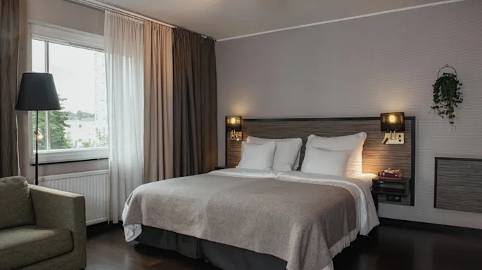 Et koselig hotellrom med en stor seng, en komfortabel lenestol ved vinduet og varm belysning, som inviterer til avslapning.