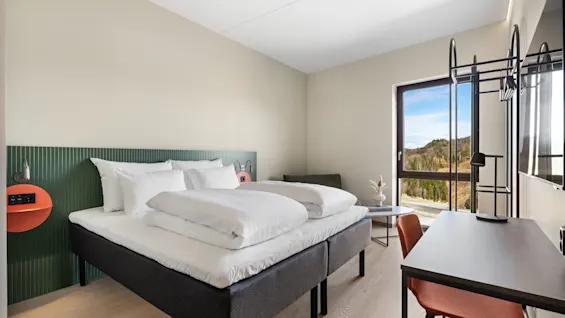 Et moderne hotellrom med to senger, et skrivebord og et stort vindu med utsikt over grønne åser og en vei.