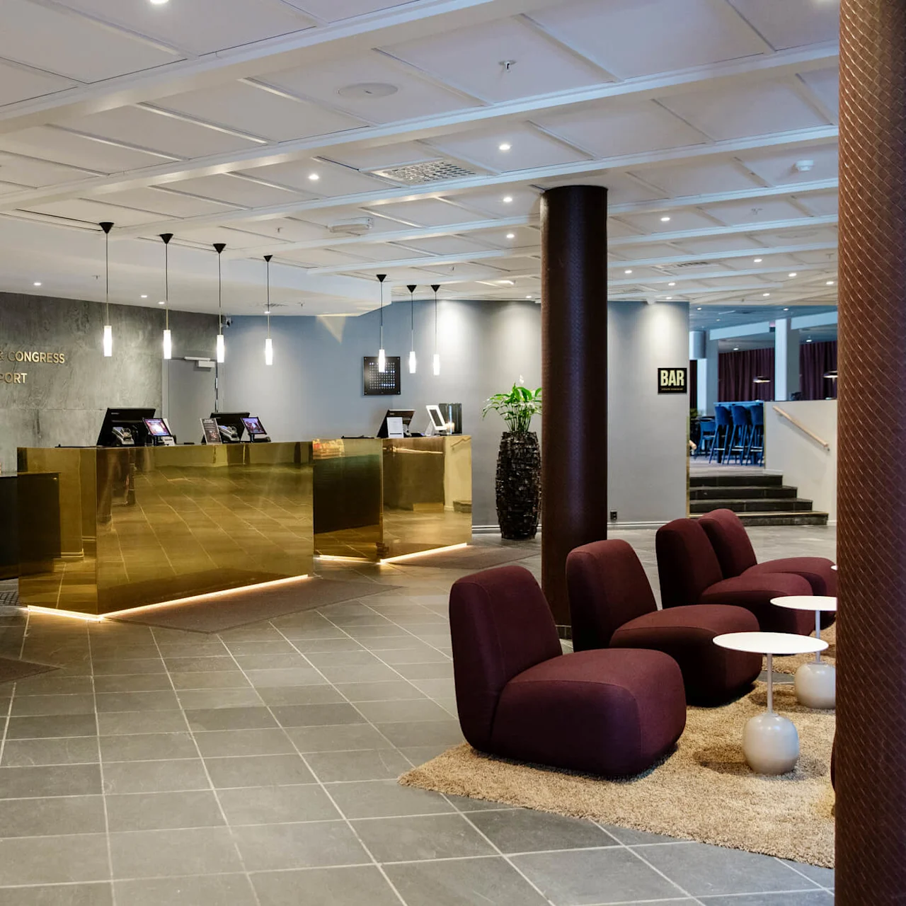 Reception med gulddetaljer står i en modern hotellobby, omgiven av lila stolar och små bord. Text lyder: "Clarion Hotel & Congress Oslo Airport."