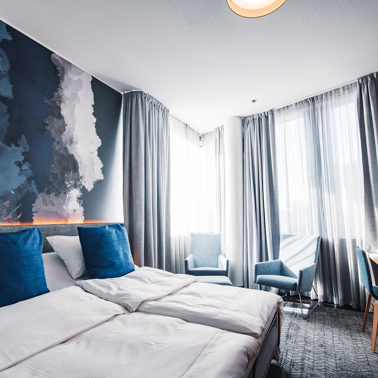A hotel room features two twin beds with blue pillows, positioned against a wall with abstract art. Two armchairs are near sunlit windows, facing a wooden desk.