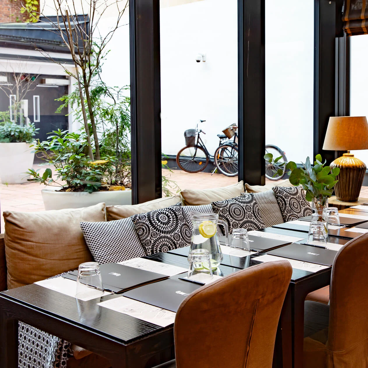 Elegant spisebord dekket med glass og kanner på en terrasse, omgitt av puter og potteplanter. En skjerm viser "Hotel Fabian". Sykler er synlige ute.