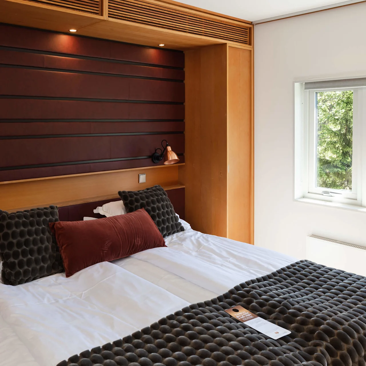 A neatly arranged bed with dark textured cushions, positioned against a wooden headboard and a window to its side, in a well-lit modern bedroom.
