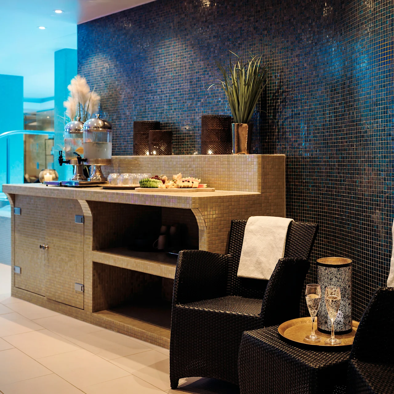A refreshment area features a countertop with tea, snacks, and greenery, surrounded by cozy chairs and robes. It is set in a spa-like environment with soft lighting.