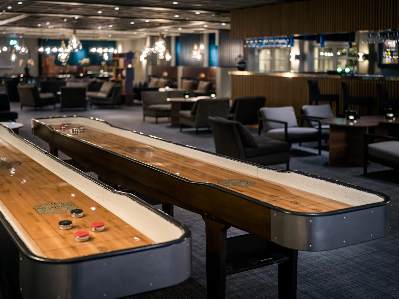 To shuffleboardbord er plassert i en moderne, dempet belyst lounge med komfortable sitteplasser og lysekroner, som skaper en avslappet og innbydende atmosfære.